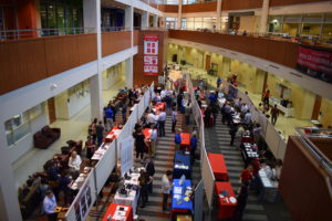 A view of most of the posters and tables set up in the HSLC atrium from above. There are three lines of poster boards, with posters set up on both sides. Many of the participants also have tables, covered in blue, red, or white table cloths.