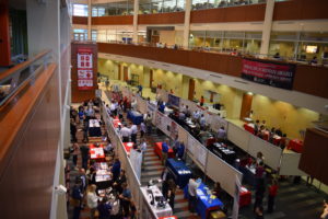 A view of most of the posters and tables set up in the HSLC atrium from above, taken at an angle. There are three lines of poster boards, with posters set up on both sides. Many of the participants also have tables, covered in blue, red, or white table cloths.