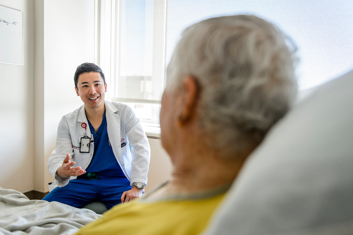A UW medical student and second year resident at the Veterans Administration Hospital, Aaron Ho, sits in front of a window, speaking to a patient sitting upright in bed.